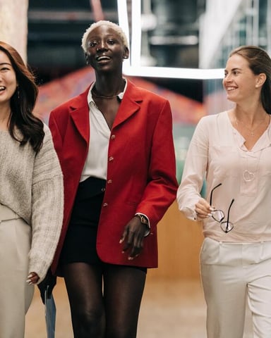 Three women walking together in a modern office hallway with brick wall and glass partitions.