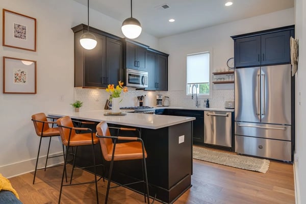 Modern kitchen with dark blue cabinetry, white subway tile backsplash, stainless steel appliances, and island with orange bar stools.