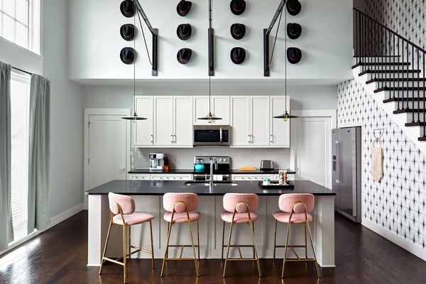 Modern kitchen with white cabinetry, black countertop island, pink bar stools, pendant lights, and decorative black hats on wall above cabinets.