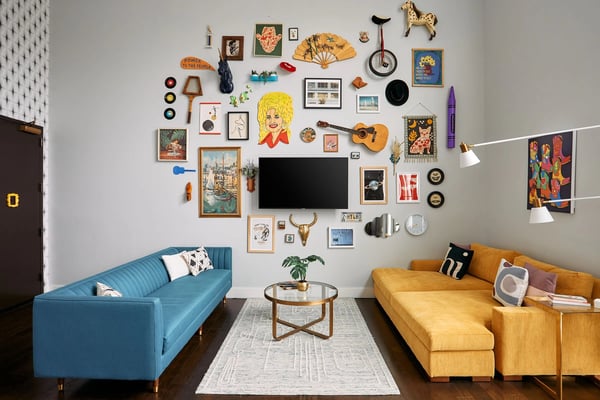 Living room with blue and yellow sofas, gallery wall of eclectic art and vintage objects, flat-screen TV, and round coffee table.