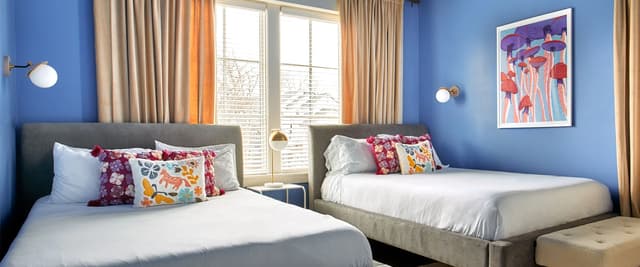 Bright blue bedroom with two queen beds, colorful patterned pillows, large windows with tan curtains, and mushroom artwork on wall.