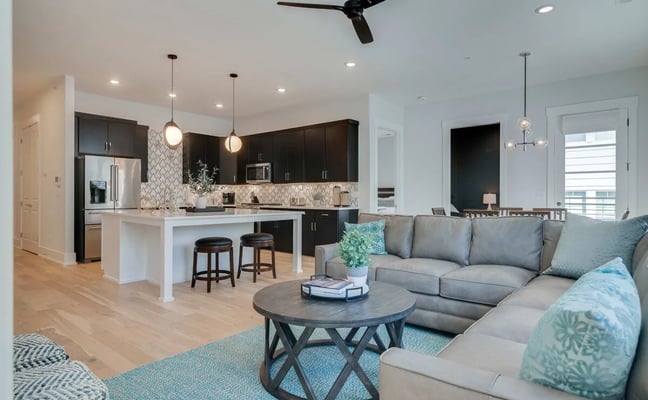 Open-plan living room and kitchen with gray sectional sofa, round coffee table, dark cabinetry, white island with bar seating, and pendant lighting.