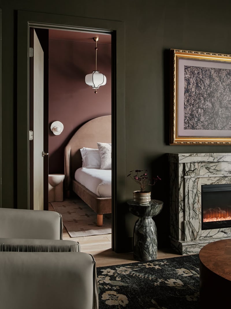 Doorway to bedroom with burgundy walls, curved headboard bed, and pendant light; fireplace with marble surround visible in foreground