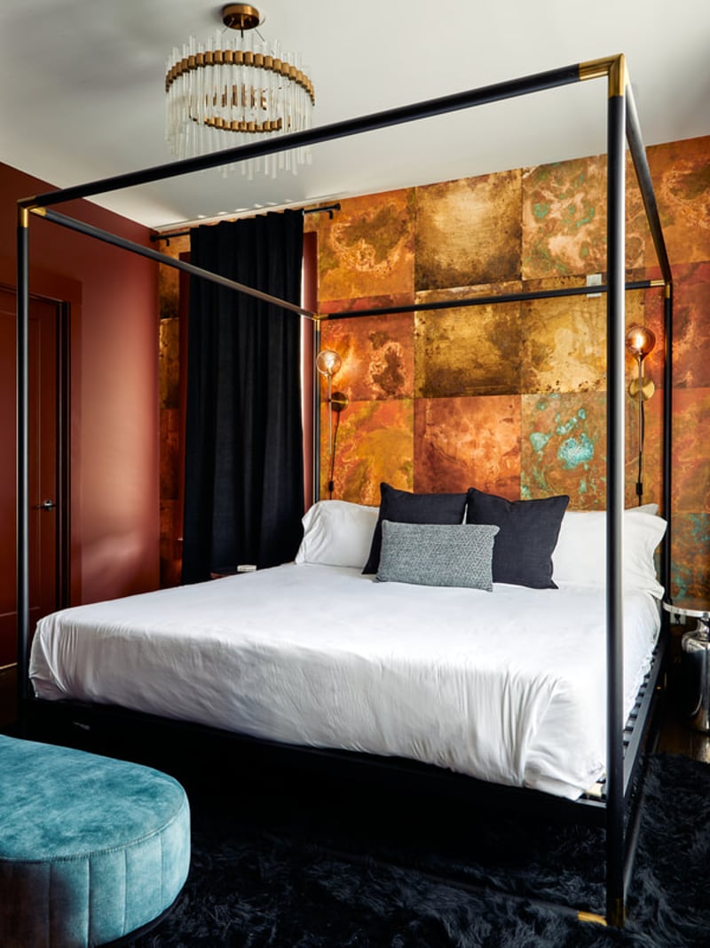 Modern bedroom with black four-poster bed, white linens, gold-leaf accent wall, brass chandelier, and turquoise ottoman