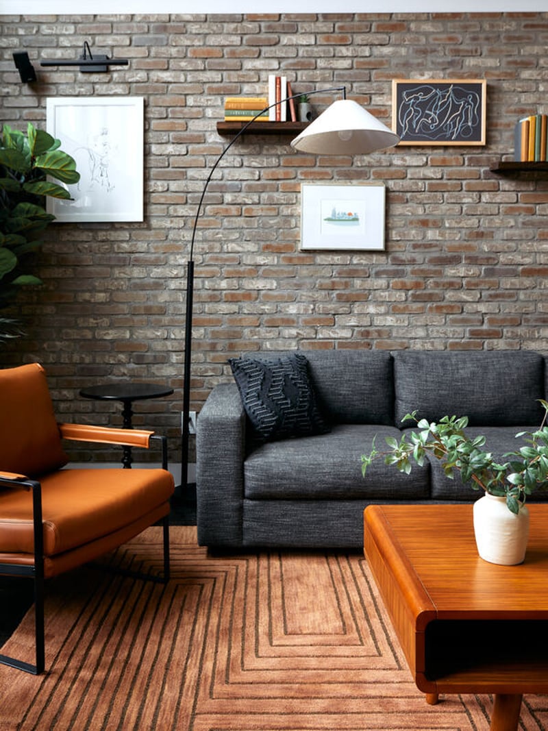 Living room with exposed brick wall, gray sofa, orange leather chair, wooden coffee table, arc floor lamp, and floating shelves with artwork and books