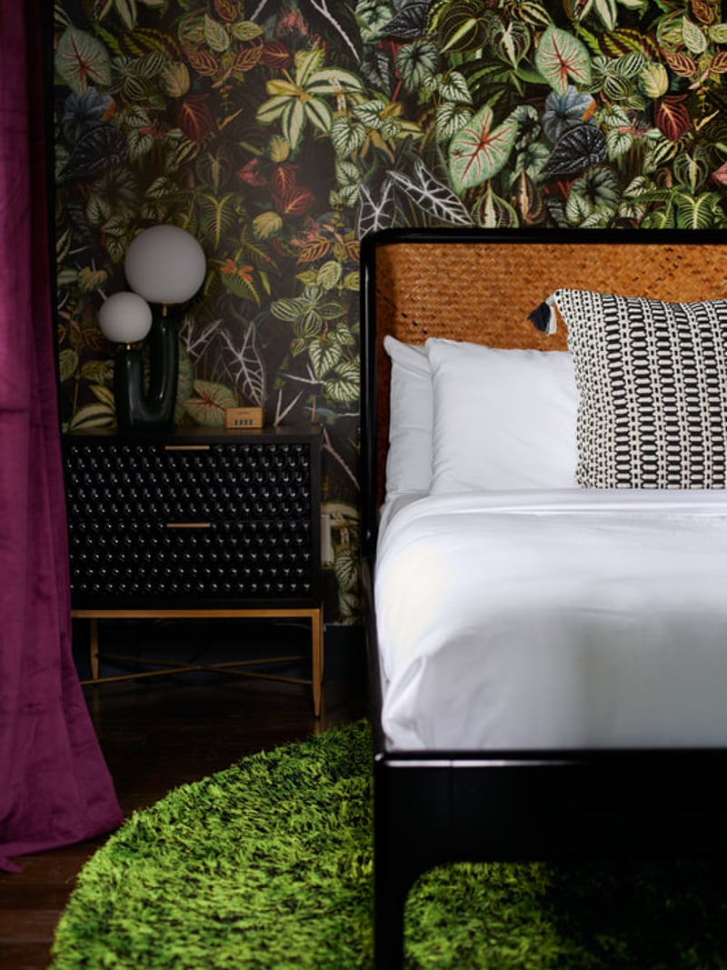 Bedroom with botanical wallpaper, black nightstand with globe lamps, bed with woven headboard and patterned pillow, bright green area rug