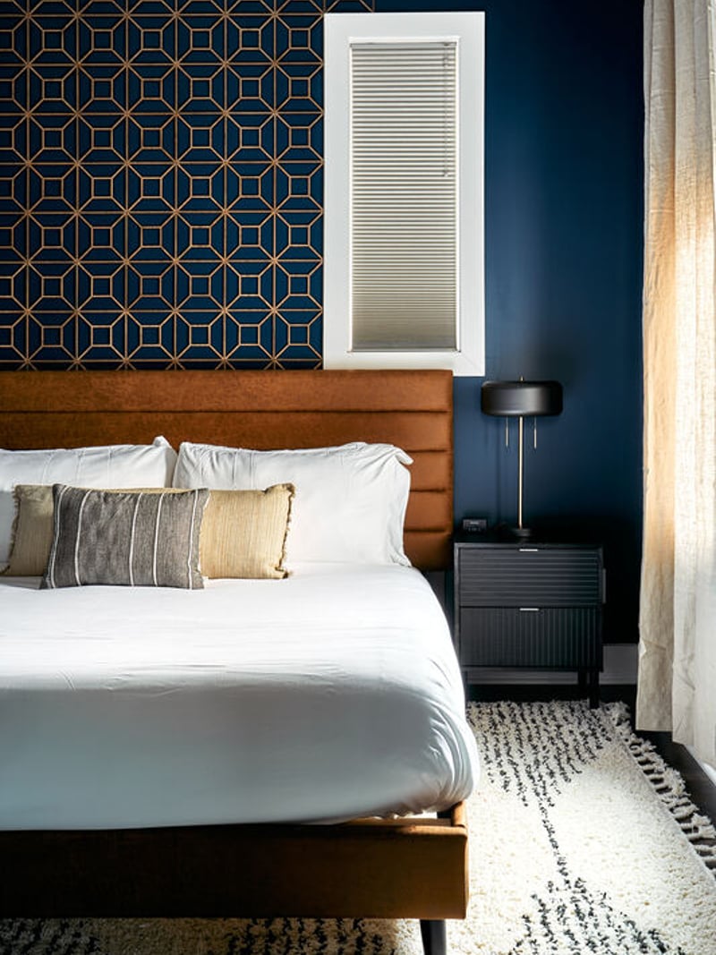 Bedroom with cognac leather headboard, white bedding, geometric patterned wallpaper, navy blue walls, and black nightstand.