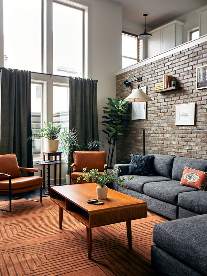 Modern living room with gray sectional sofa, orange accent chair, wood coffee table, exposed brick wall, and tall windows with dark curtains