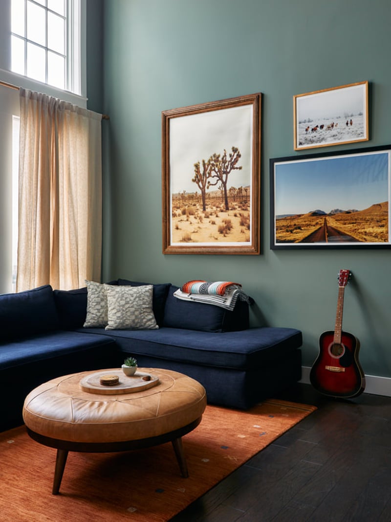 Living room with blue sofa, round leather ottoman, desert landscape artwork, acoustic guitar, and large window with sheer curtains