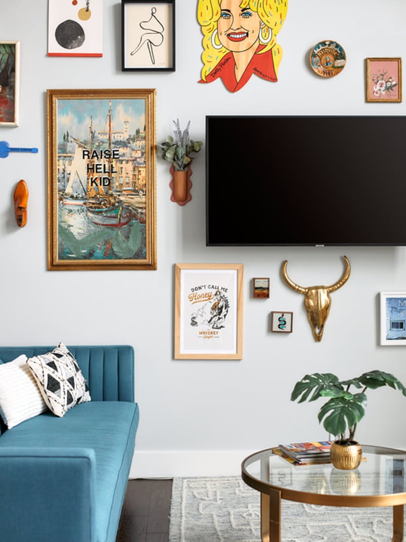 Living room with blue sofa, gallery wall of eclectic artwork including portrait and vintage prints, flat-screen TV, and round coffee table with plant.