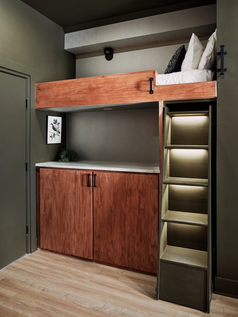 Loft bed with wooden frame, white bedding, and built-in storage cabinets below with illuminated shelving unit on the side
