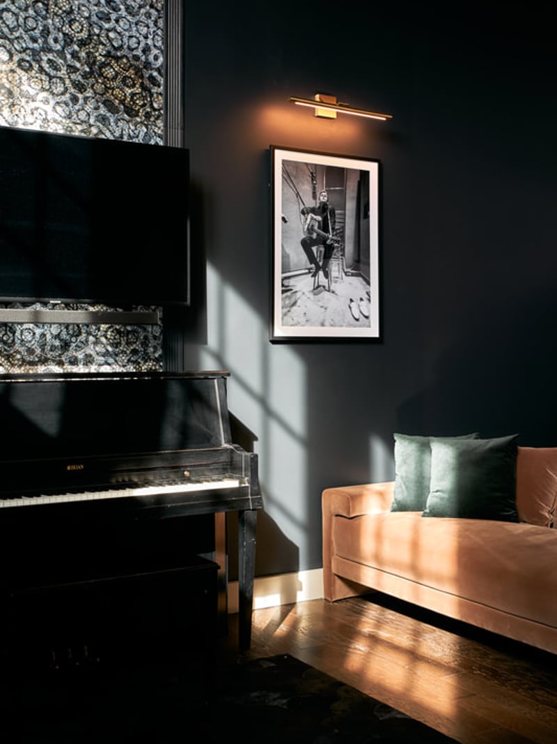 Dark living room with black piano, tan sofa with green pillows, framed black-and-white photograph, and brass wall sconce