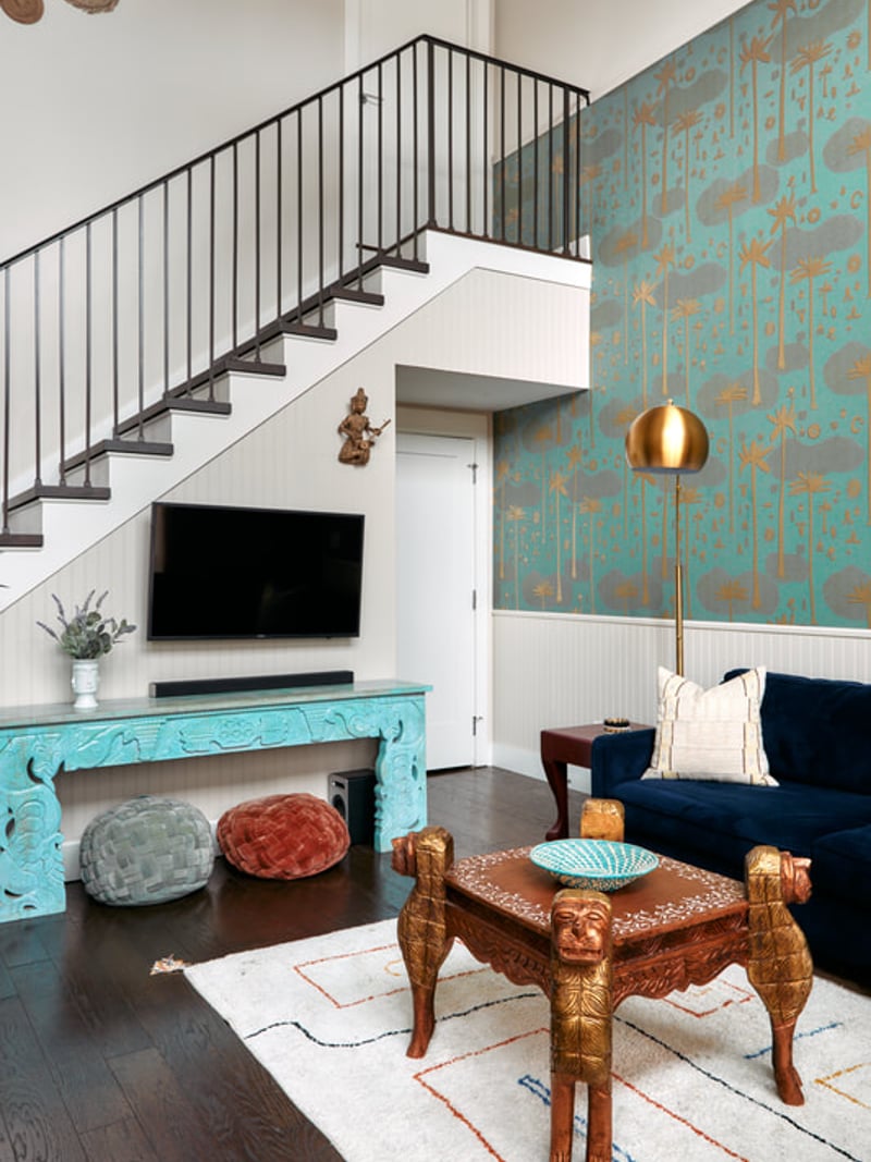 Modern living room with turquoise patterned wallpaper, wooden carved bench, navy sofa, and white staircase with black railing overhead.