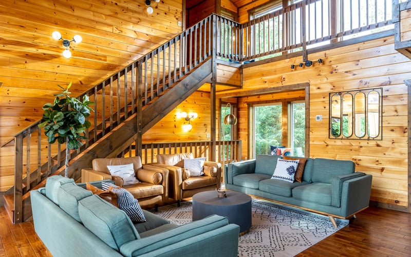 Spacious wooden cabin interior with two-story loft, multiple seating areas, metal staircase, and large windows overlooking trees