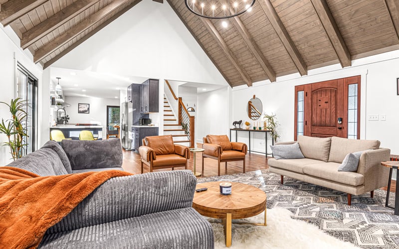 Open-plan living room with vaulted wood ceiling, gray and orange seating, wooden staircase, and red front door