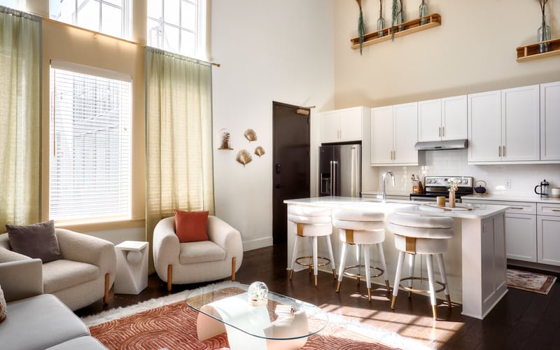 Open-plan living room and kitchen with white cabinetry, island seating, cream furniture, and large windows with green curtains