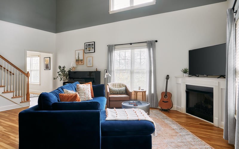 Open-plan living room with blue sectional sofa, fireplace with TV, acoustic guitar, and wooden staircase visible on left