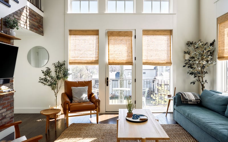 Bright living room with high ceilings, three glass doors with woven shades, tan armchair, blue sofa, and fireplace with brick surround.