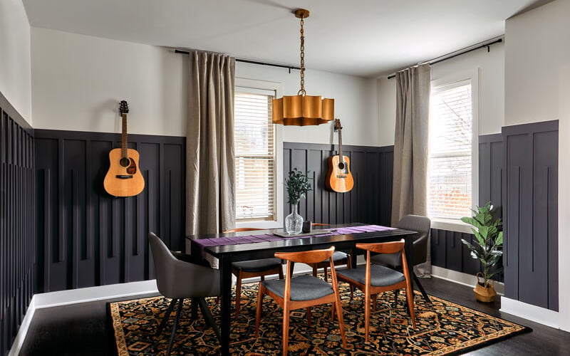 Dining room with black shiplap walls, wooden chairs, gold pendant light, two acoustic guitars, and patterned area rug.