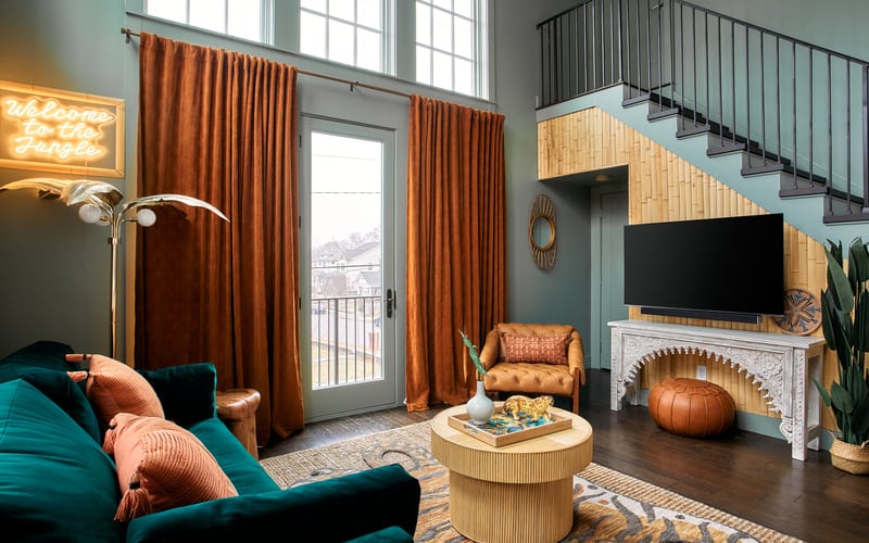 Modern loft living room with teal sofa, orange curtains, fireplace with TV, and metal staircase.
