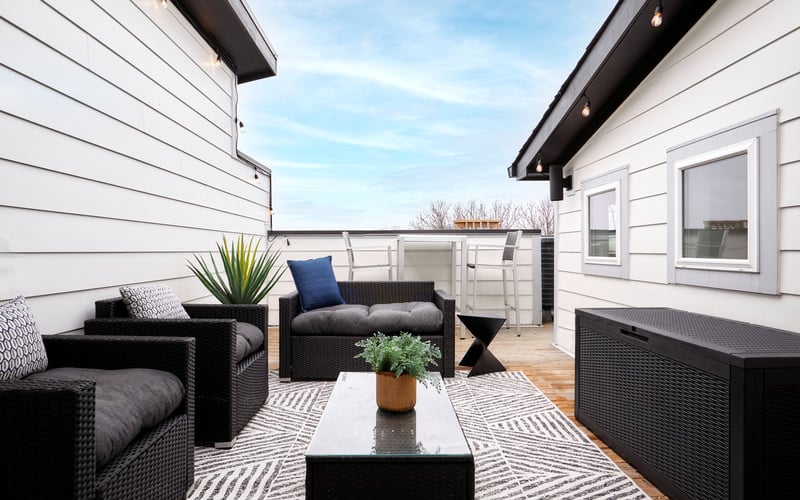 Modern deck with black wicker furniture, white and gray cushions, potted plant, and clear sky view between white-sided buildings