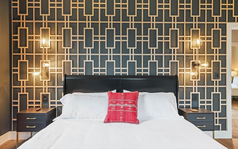 Bedroom with geometric patterned wallpaper, black bed frame, white bedding, red accent pillow, and brass wall sconces