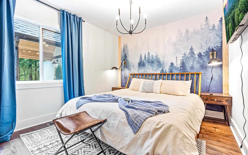 Bedroom with wooden bed, forest mural, blue curtains, pendant light, and window overlooking trees
