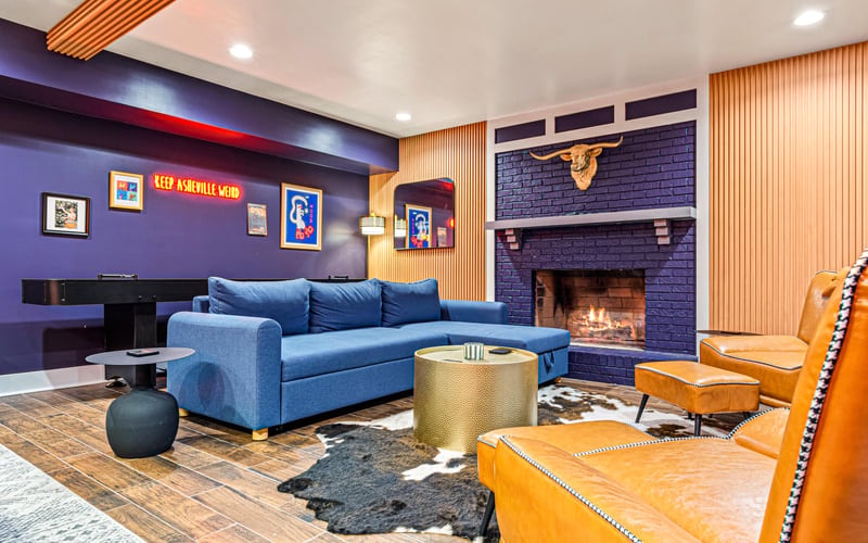 Colorful basement lounge with blue sectional sofa, fireplace, orange leather chairs, and neon signage on purple walls