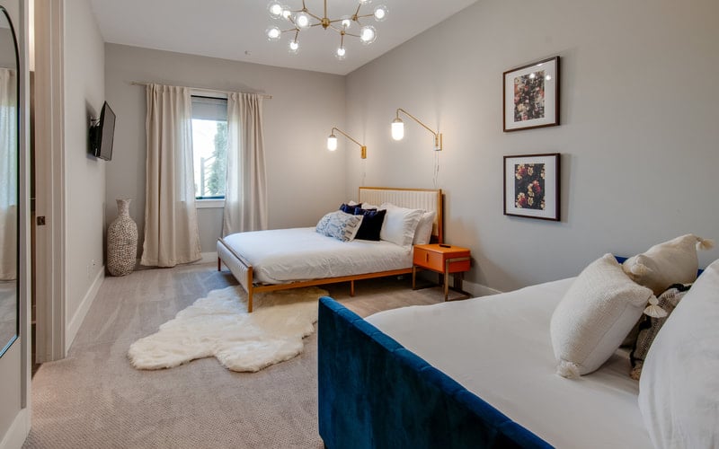 Modern bedroom with two beds, wooden nightstand, pendant lighting, framed artwork, and large window with curtains