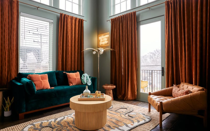 Modern living room with teal velvet sofa, cognac leather chair, wooden round table, rust curtains, and balcony doors with winter view