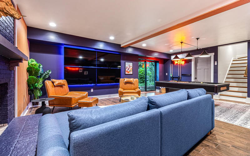 Modern basement entertainment room with blue sectional sofa, orange leather chairs, large wall-mounted screens, and neon signage