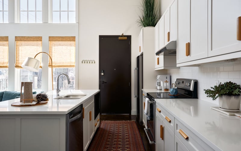 Modern galley kitchen with white cabinetry, black appliances, white countertops, and wood-framed windows with natural light