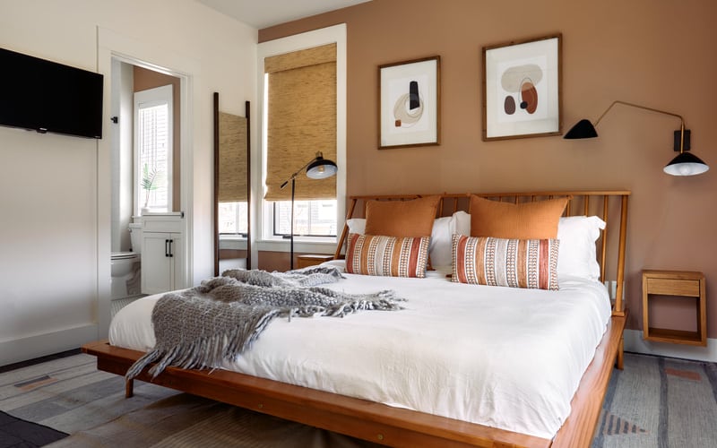 Bedroom with wood platform bed, white bedding, orange and striped pillows, terracotta accent wall, and ensuite bathroom visible through open door