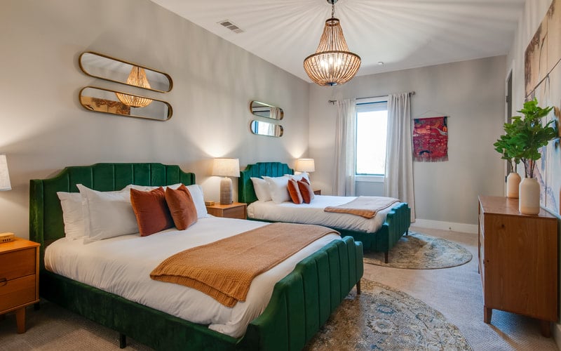 Bright bedroom with two green velvet beds, pendant light fixture, white walls, and wood furnishings