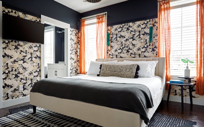 Bedroom with cream upholstered bed, navy and cream floral wallpaper, orange curtains, and dark wood flooring