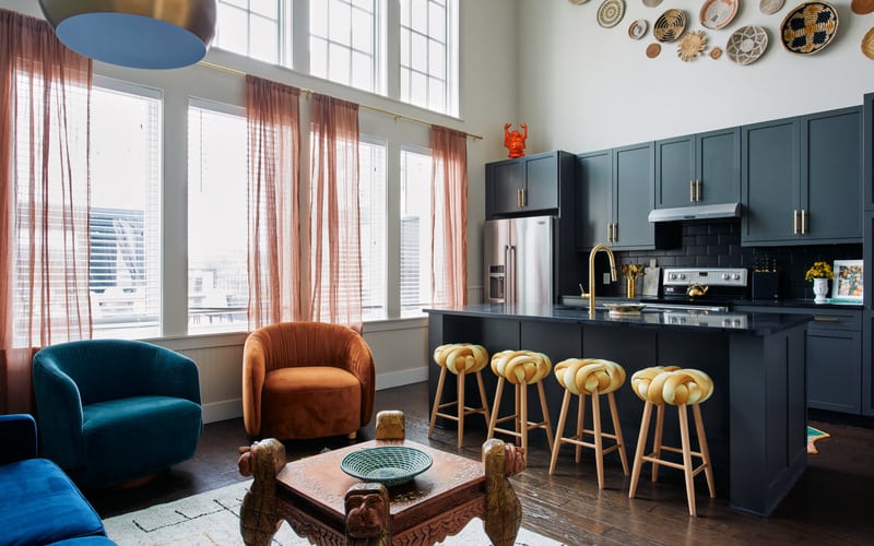Modern open-plan kitchen and living room with dark blue cabinetry, island seating, teal and rust velvet chairs, and floor-to-ceiling windows with red plaid