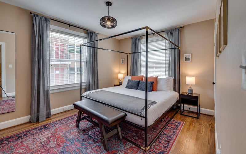 Bedroom with black four-poster bed, hardwood floors, red patterned rug, and large window with gray curtains