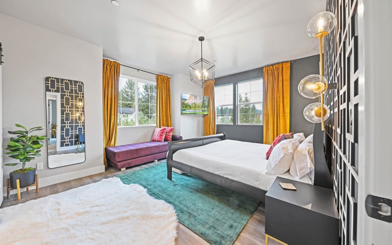 Modern bedroom with queen bed, burgundy sofa, gold curtains, pendant lights, and views of trees through large windows