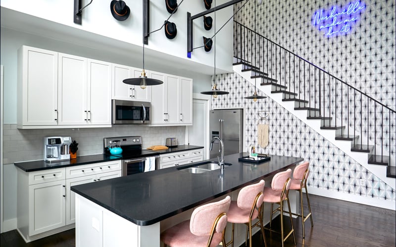 Modern kitchen with white cabinetry, black countertops, island seating with pink velvet chairs, and staircase with neon sign.