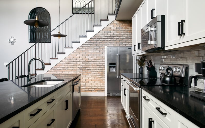 Modern kitchen with white cabinetry, black countertops, stainless steel appliances, and exposed brick wall leading to staircase with black metal railings.