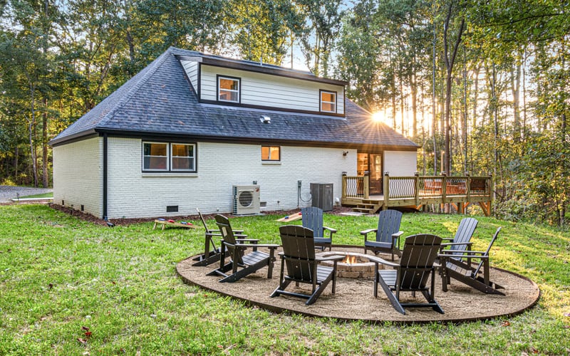 White brick house with dark roof surrounded by trees, fire pit seating area with Adirondack chairs in foreground at sunset.