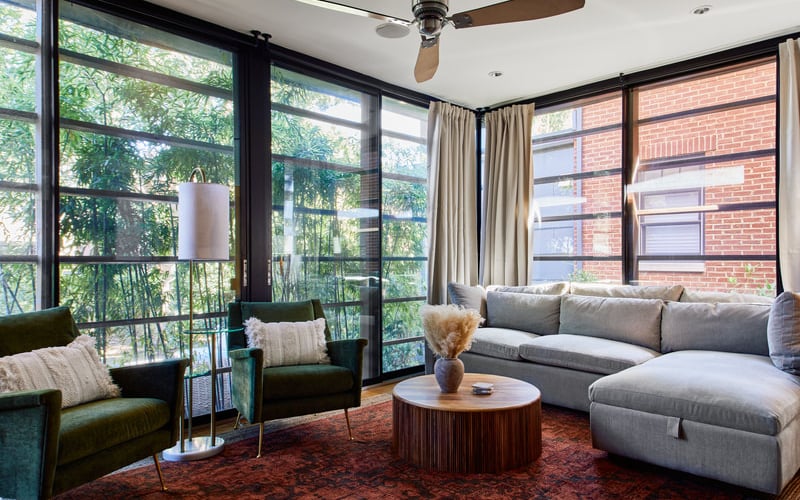 Modern living room with gray sectional sofa, green armchairs, round wooden coffee table, and floor-to-ceiling windows overlooking trees and brick building.