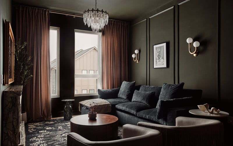 Dark moody living room with black sofa, round coffee table, crystal chandelier, and floor-to-ceiling windows with brown curtains
