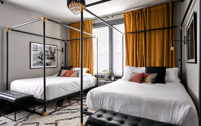 Modern bedroom with two four-poster beds, gold curtains, crystal chandelier, and framed artwork on walls