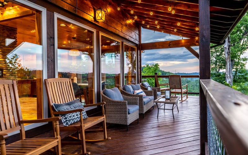 Covered deck with wooden beams, string lights, Adirondack chairs, wicker seating, and panoramic valley views at dusk