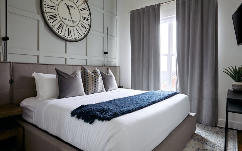 Modern bedroom with gray upholstered bed, white bedding, decorative pillows, large wall clock, gray curtains, and potted plant
