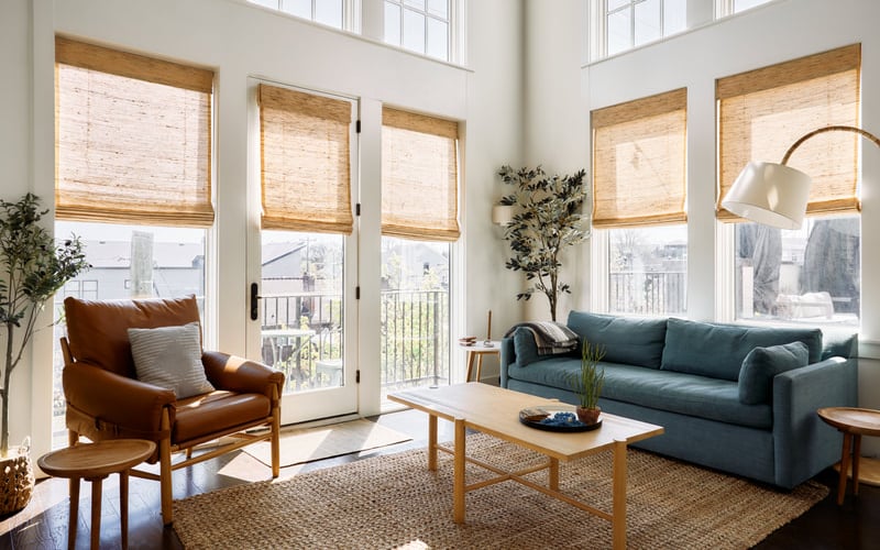 Bright living room with blue sofa, cognac leather chair, wooden coffee table, and multiple windows with woven shades overlooking city skyline.