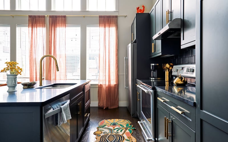 Modern kitchen with black cabinetry, white countertops, gold fixtures, and coral sheer curtains on tall windows