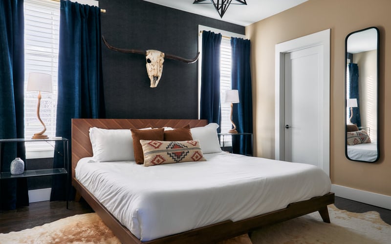 Modern bedroom with wood bed frame, white bedding, dark accent wall with animal skull, blue curtains, and tan walls