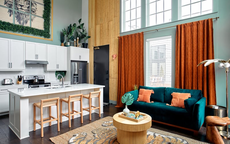 Open-plan kitchen and living room with white cabinetry, teal walls, teal sofa with orange pillows, and orange curtains